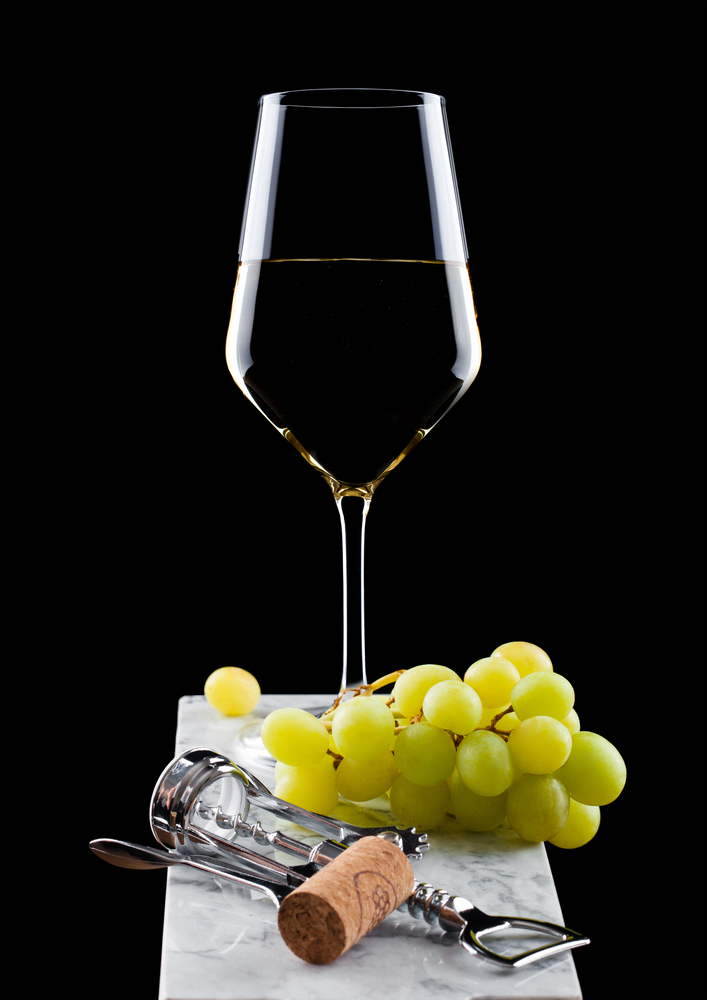 Glass of white wine with green grapes on marble board with corkscrew opener and cork on black background.