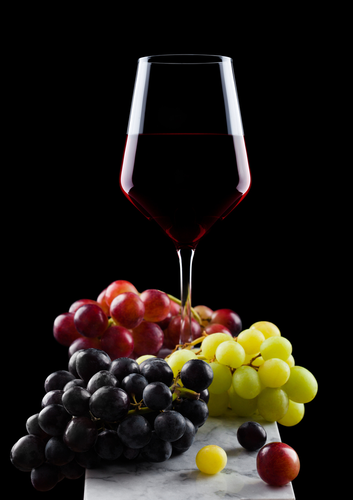 Elegant glass of red wine with dark and red and green grapes on wooden board on black background.
