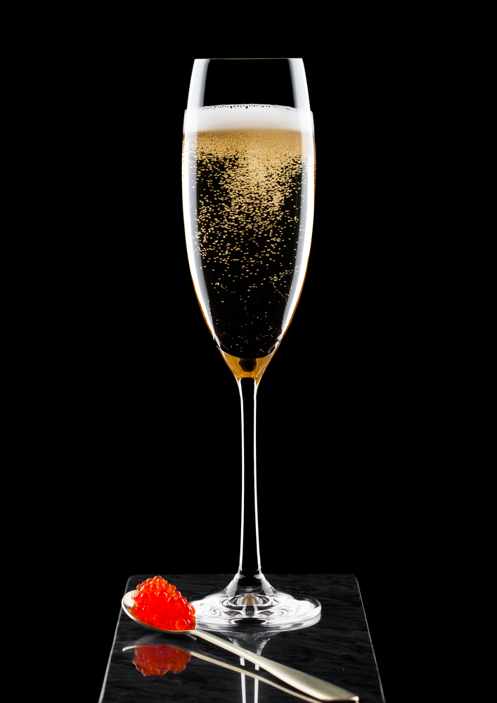 Elegant glass of yellow champagne with red caviar on golden spoon of caviar on marble board on black.