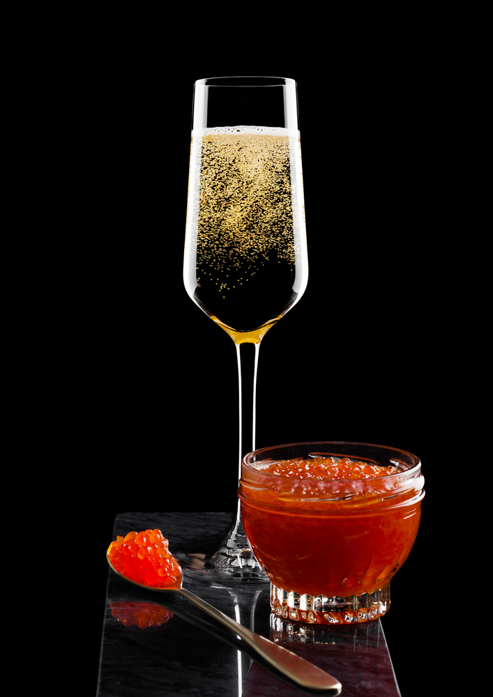 Elegant glass of yellow champagne with red caviar on golden spoon and glass container of caviar on marble board on black.