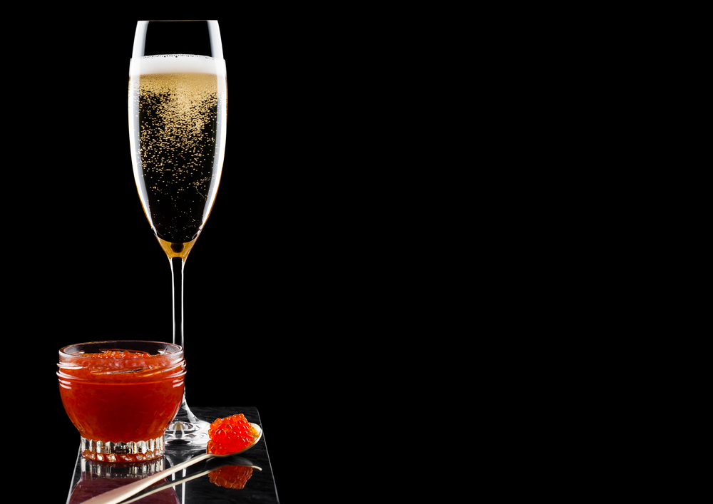 Elegant glass of yellow champagne with red caviar on golden spoon and glass container of caviar on marble board on black background