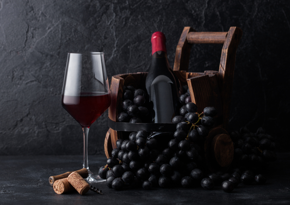 Elegant glass of red wine with dark grapes and bottle of wine inside vintage wooden barrel on black stone background.