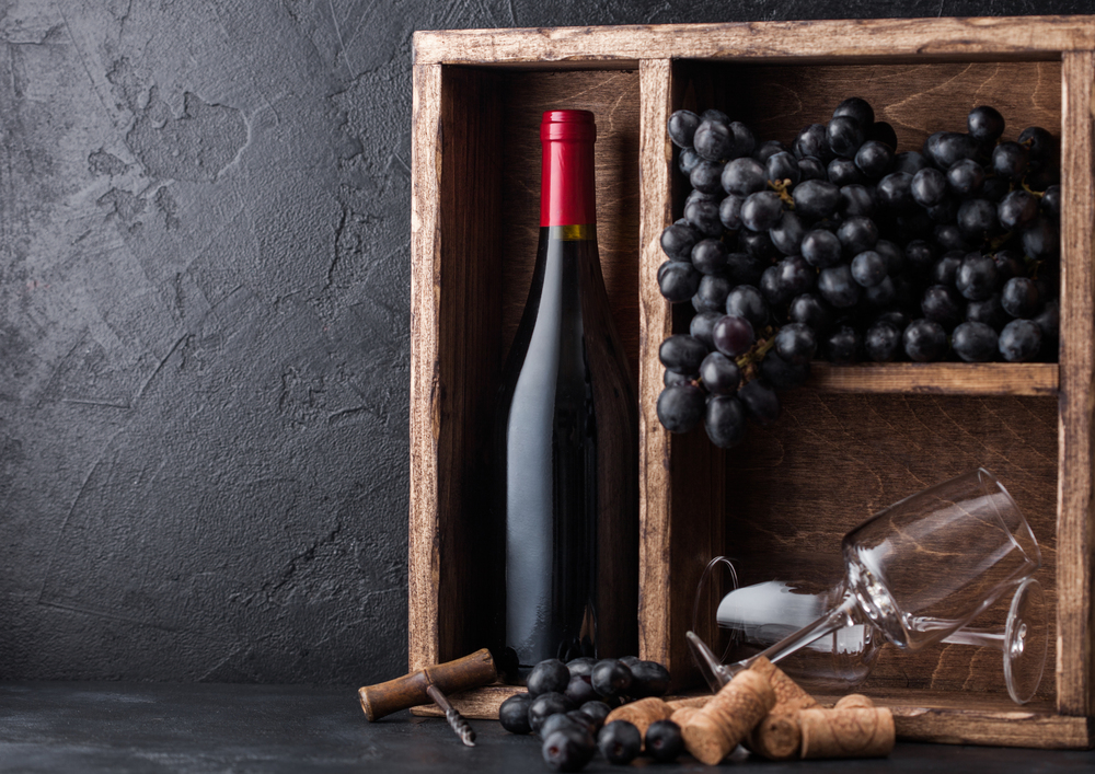 Bottle of red wine and empty glasses with dark grapes inside vintage wooden box on black stone background. Corks and corkscrew on black board.