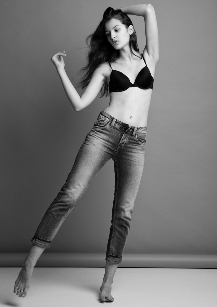 Model test portrait with young beautiful fashion model posing on grey background. Black bra and jeans