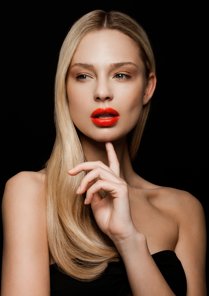 Beauty fashion model portrait with shiny blonde hairstyle with red lips on black background holding finger next to her chin
