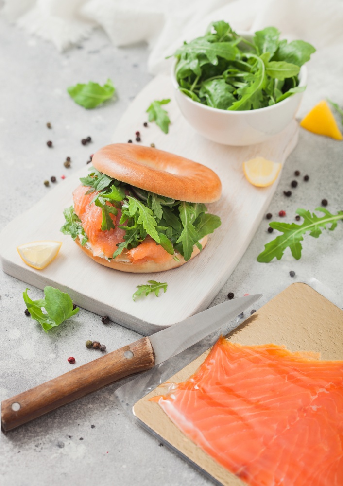 Fresh sandwich with bagel and salmon, cream cheese and wild rocket on white board with smoked salmon pack and knife on light background.