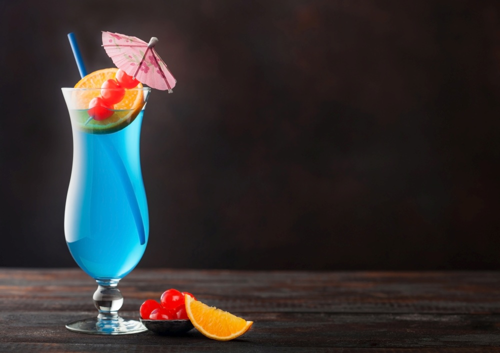 Blue lagoon summer cocktail in classic glass with sweet cocktail cherries and orange slice with umbrella on dark table background. Space for text