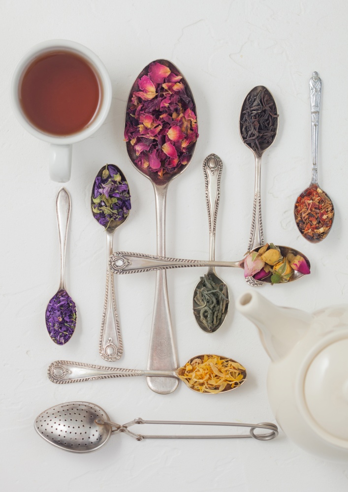 Various spoons with black and green tea, rose buds mix and blue mallow flowers and petals on white background with teapot and cup. Top view