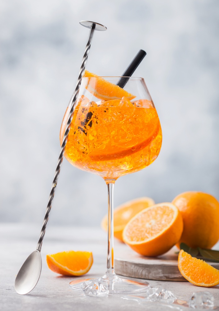 Glass of aperol spritz summer cocktail with oranges and bar spoon on light table background. Ice cubes and black straw.