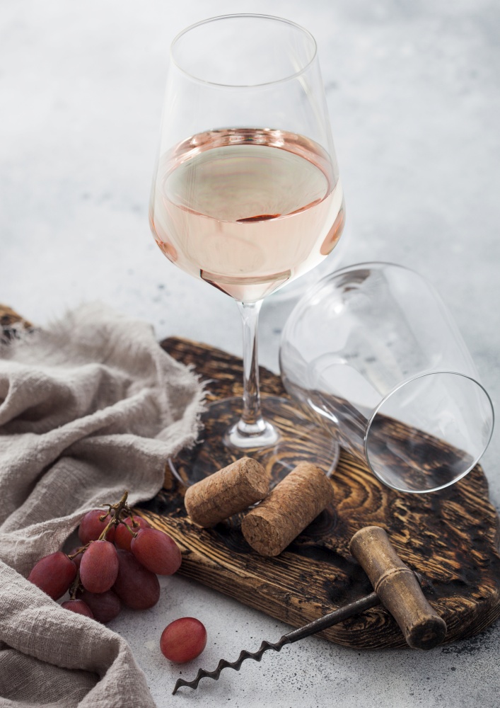 Glass of rose pink homemade wine with corks, corkscrew and grapes on wooden board with linen cloth on light table background.