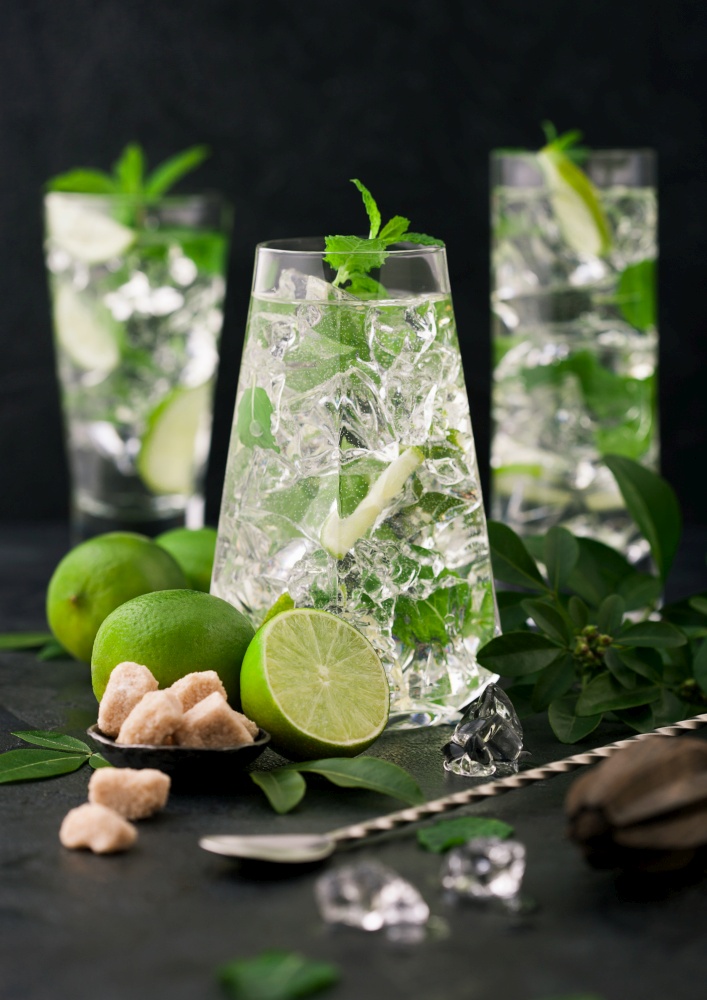 Glasses of Mojito cocktail with ice cubes,mint and lime on black board with spoon and fresh limes with cane sugar. Best party drink.Sparkling mint water