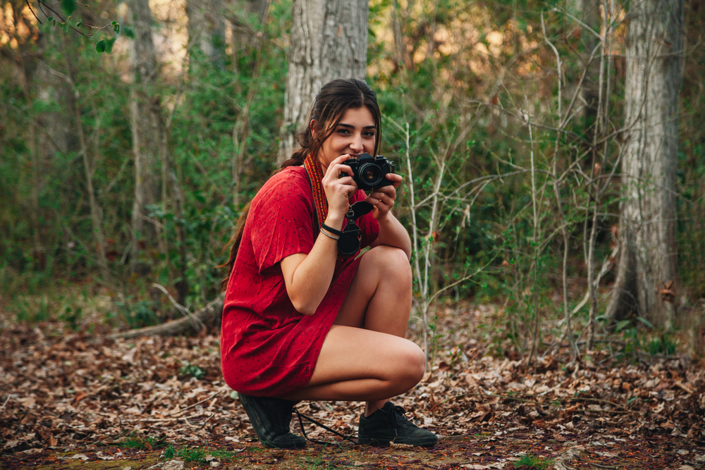 Young smiling woman taking photos in the forest with her analogical camera wearing a red mini dress.