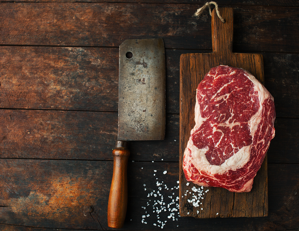Raw fresh ribeye steak with salt and an ax on a wooden background