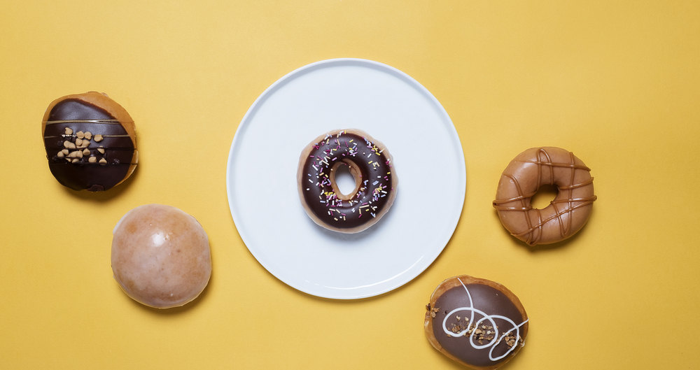 Chocolate donut, doughnut, on a white plate surrounded by various donuts on a yellow background