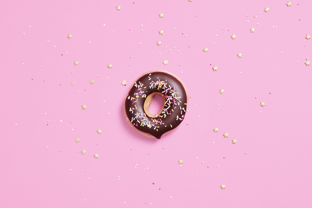 Overhead shot of single donut, doughnut, with chocolate glaze and sprinkles on a pink background with gold coloured star sprinkles scattered around it with copy space and room for text