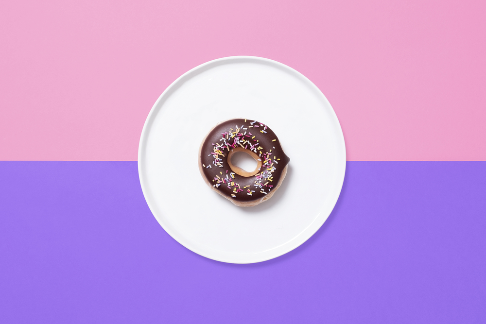 Overhead shot of single chocolate donut, doughnut, with sprinkles on a white plate on a split pink and purple background with star and heart shaped sprinkles scattered