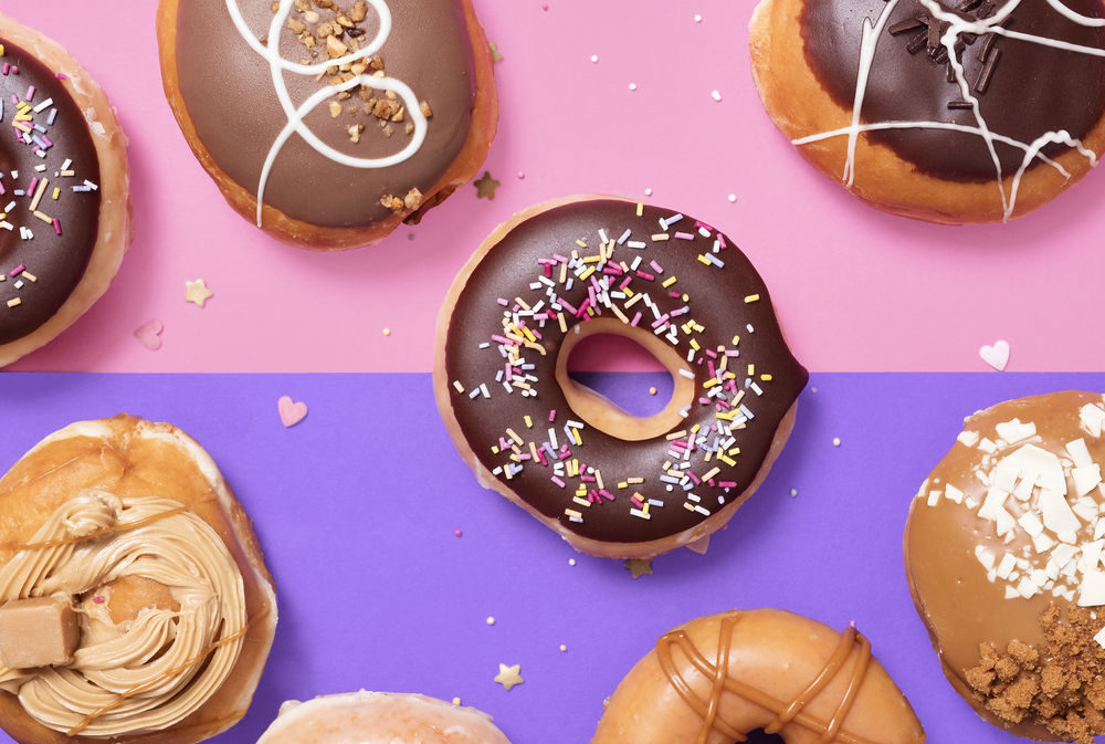 Overhead shot of donuts, doughnuts, on a split colour background of pink and purple with scattered heart and star sprinkles with chocolate and glaze and swirls