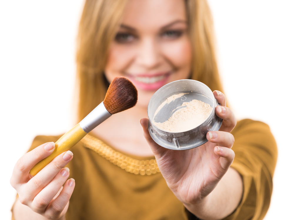 Fashionable cheerful young female holding professional powder brush, adding last touch to her make up. Smiling woman applying face powder