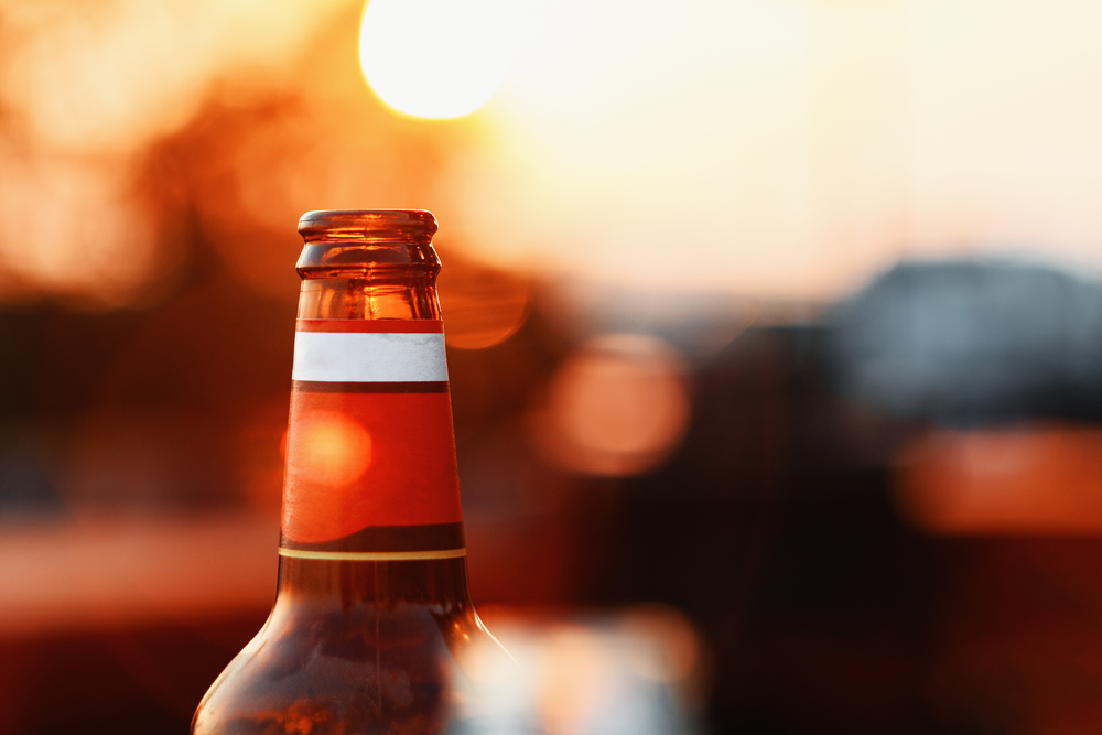 Beer Bottle Against Summer Sky At Sunset On Blurred Background With Lens Flare