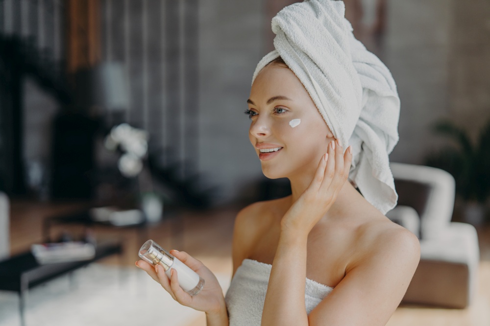 Beautiful young European woman with white bath towel on head, applies face cream and body lotion, enjoys skin care procedures after shower, poses at home alone. Daily beauty routines concept