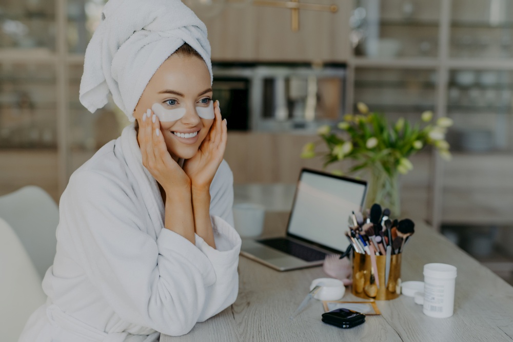 Pretty smiling young female model applies collagen patches under eyes to reduce wrinkles wears bathrobe and towel on head enjoys skin care procedures sits at desk with cosmetic tools cozy interior