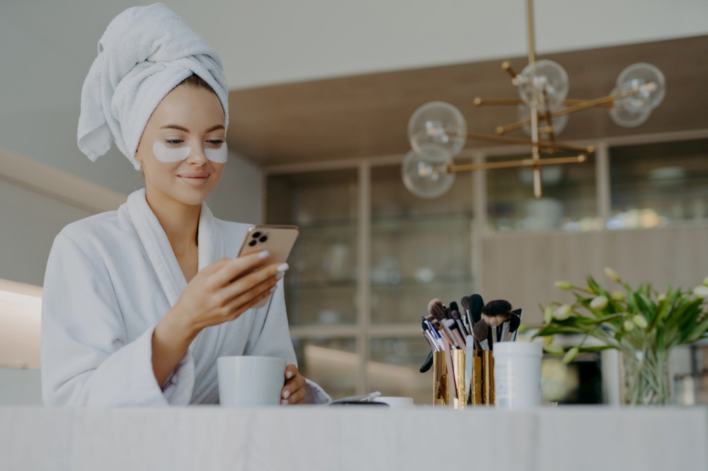 Photo of attractive young woman wears bath towel on head and soft white dressing gown holds mobile phone enjoys online communication applies moisturising undereye patches drinks hot beverage