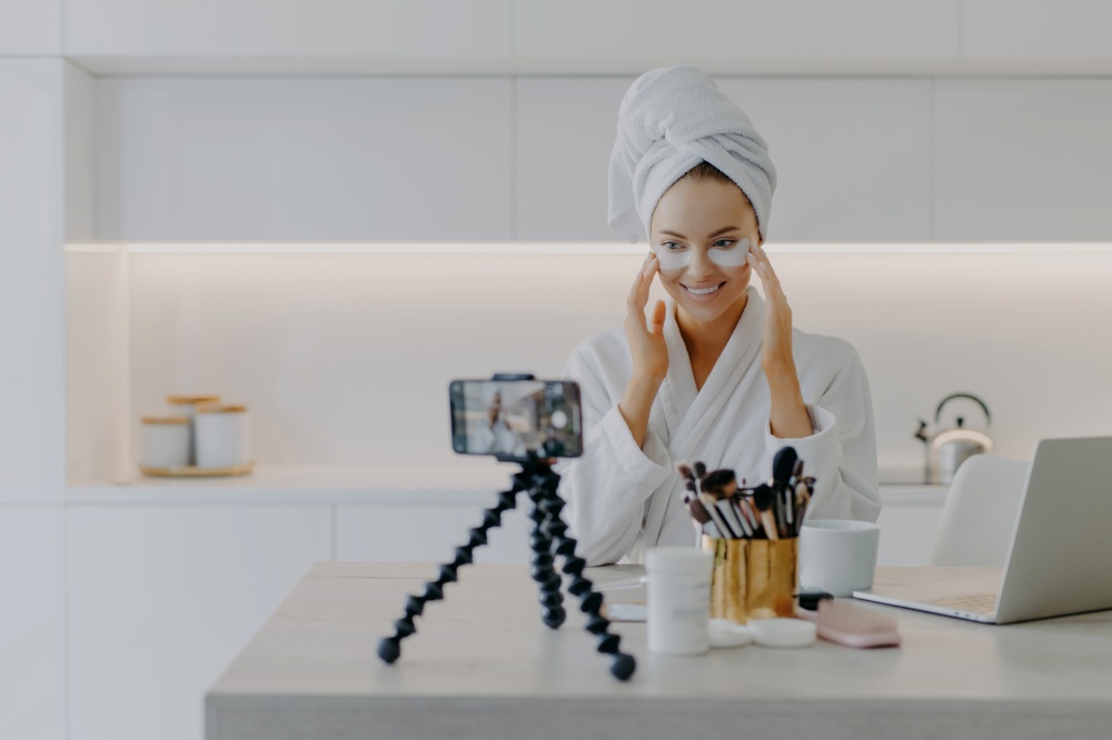 Happy young Caucasian woman records beauty blog on mobile phone standing on tripod goves advice how to use cosmetic patches shoots video wears bathrobe poses at home uses high speed internet