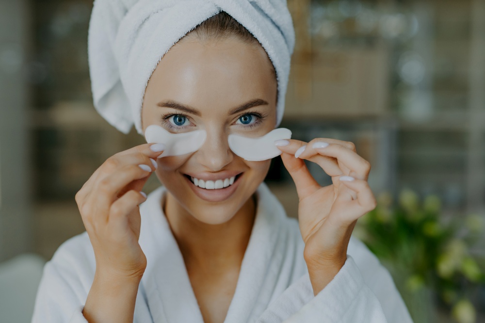 Headshot of happy young blue eyed woman with minimal makeup takes off beauty patches enjoys good effect has healthy well cared skin white perfect teeth wears bathrobe and wrapped towel on head