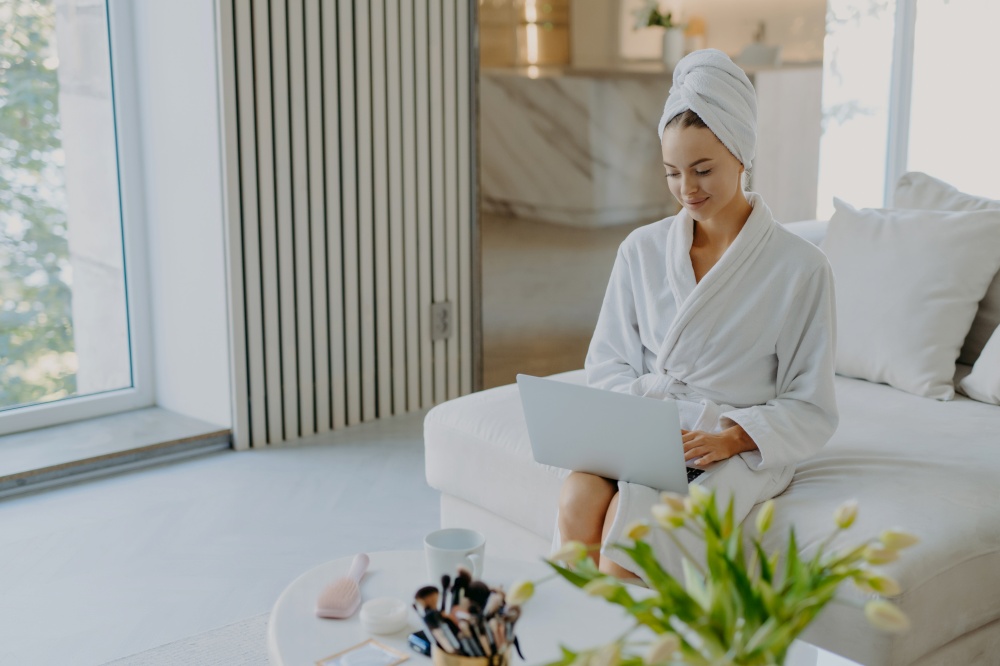 Photo of attractive female model wears bathrobe and towel types on laptop computer works freelance sits on comfortable sofa enjoys domestic atmosphere poses against modern apartment interior