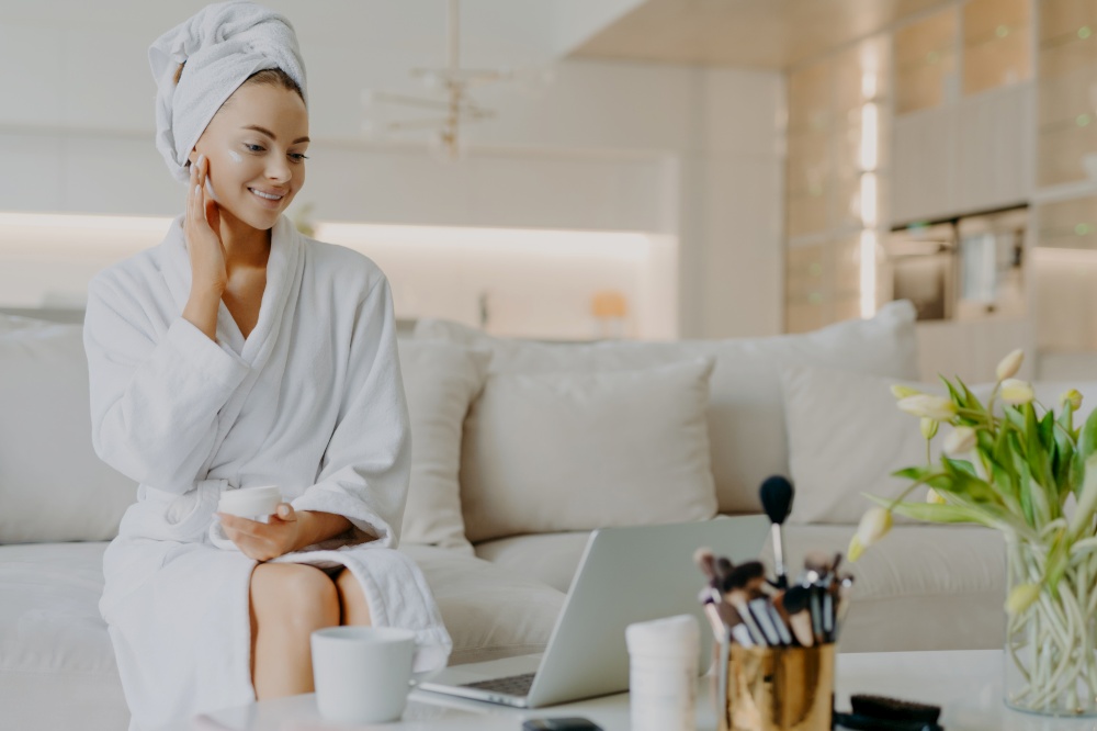 Photo of smiling woman applies nourishing cream on face looks in laptop screen gives advice of skin care dressed in bathrobe and towel sits at comfortable sofa has rejuvenation procedure. Cosmetology