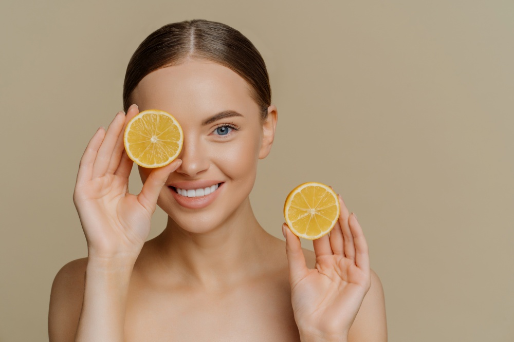 Isolated shot of good looking tender brunette woman smiles gently at camera covers eye with half of lemon holds favorite fruit poses topless indoor against beige background. Skin care concept