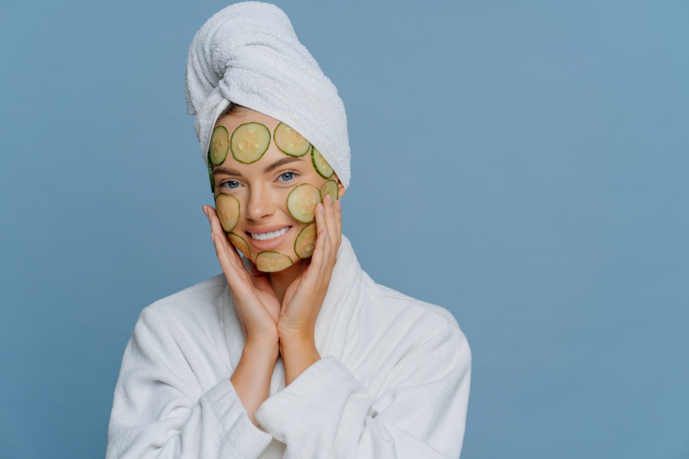 Skin care and beauty treatment concept. Happy relaxed young female model applies cucumber slices to nourish skin keeps hands on face dressed in white dressing gown and wrapped towel on head.