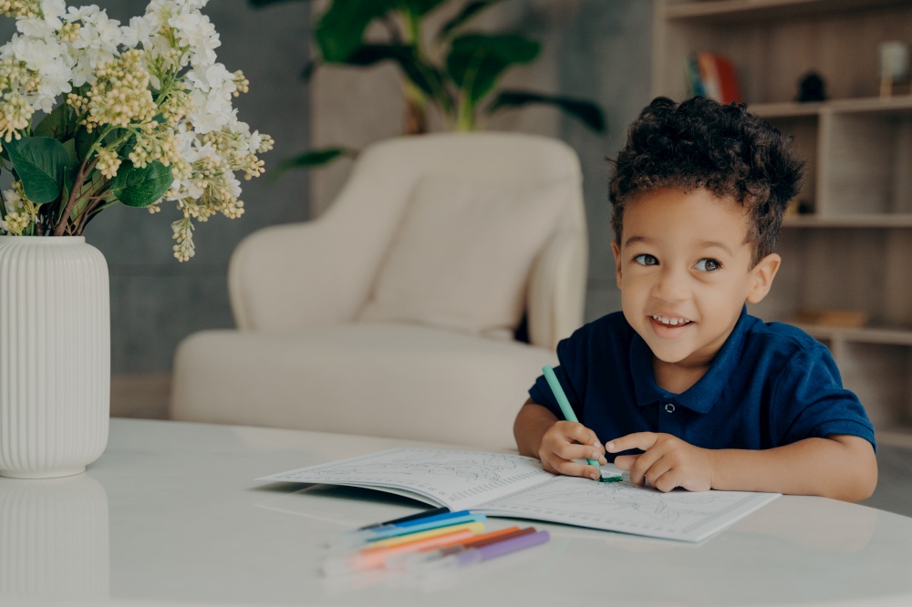 Happy curly mixed race boy coloring animals in coloring book with felt pens, looking aside and smiling while sitting in front of table in comfy living room at home. Children leisure time activities. Cute afro american child painting in coloring book at home