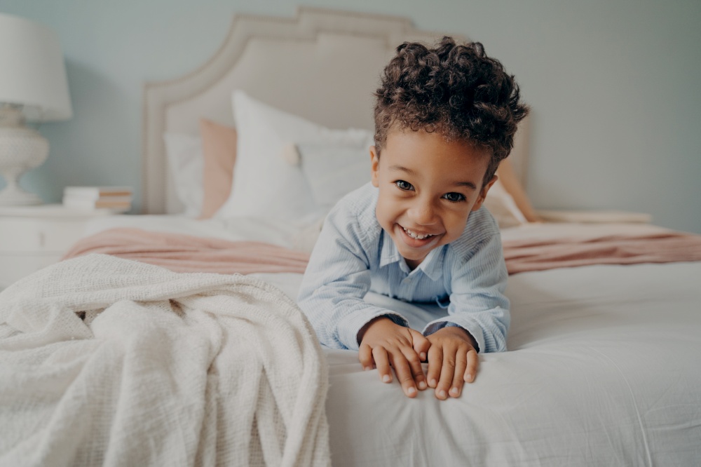 Portrait of happy curly mulatto boy smiling at camera while laying on cozy bed, relaxing after fun playing session at home, adorable preschool child enjoying indoor activities and leisure time. Happy curly afro american boy laying on bed and smiling at camera