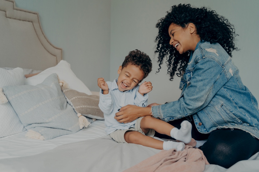 Happy mixed race family at home, carefree young woman mom in denim jacket and jeans with curly hair playing with her sweet child little boy, tickling his tummy while spending time together in bedroom. African american woman having fun with little adorable son at home
