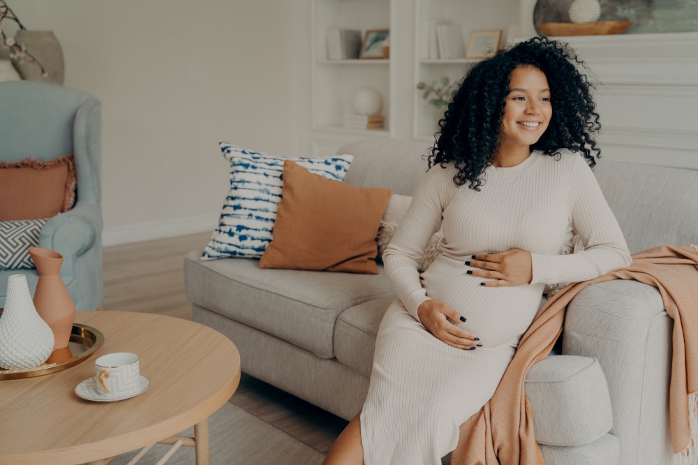 Happy pregnant afro american woman in casual dress with curly hair looking at window while sitting on couch in living room, checking out weather and thinking about going outside for walk. Pregnant afro american woman looking at window while sitting on sofa at home