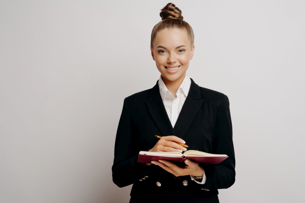 Female lawyer or business owner in dark suit with classic hairstyle writing down information about clients in red notebook with pencil while standing against light colored wall, smiling at camera. Business woman in dark suit writing in note book