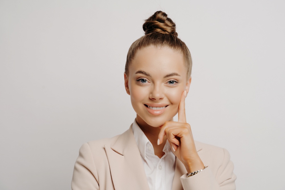 Woman office worker in beige suit with hair in bun looking happily at camera, doing confident gesture and thinking about promotion and career while posing against grey background. Office worker in beige suit feeling happy