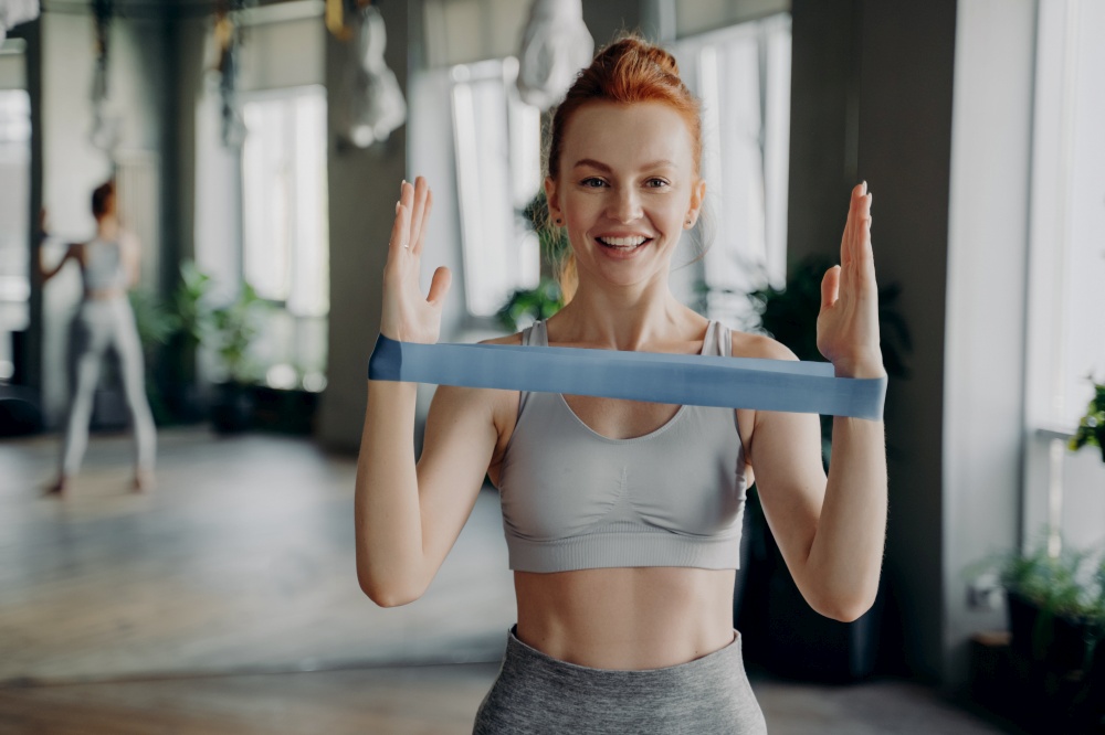 Portrait of young sporty happy redhead woman wearing white top and gray leggings performs exercises for muscles of hands during workout with resistance band. Fitness instructor during sports class. Portrait of young sporty redhead woman in sportswear with resistance rubber band