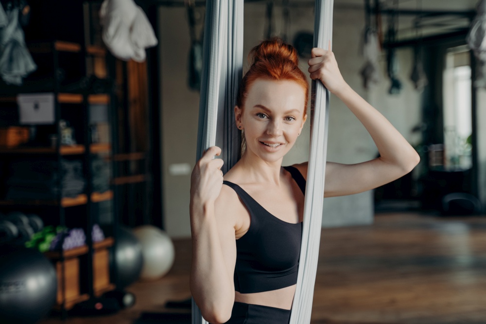Portrait of young active and healthy female in black top sitting on fly yoga hammock and hanging over floor in gym during sports class in fitness studio background, relaxing and smiling at camera. Young active female in tracksuit sitting on fly yoga hammock