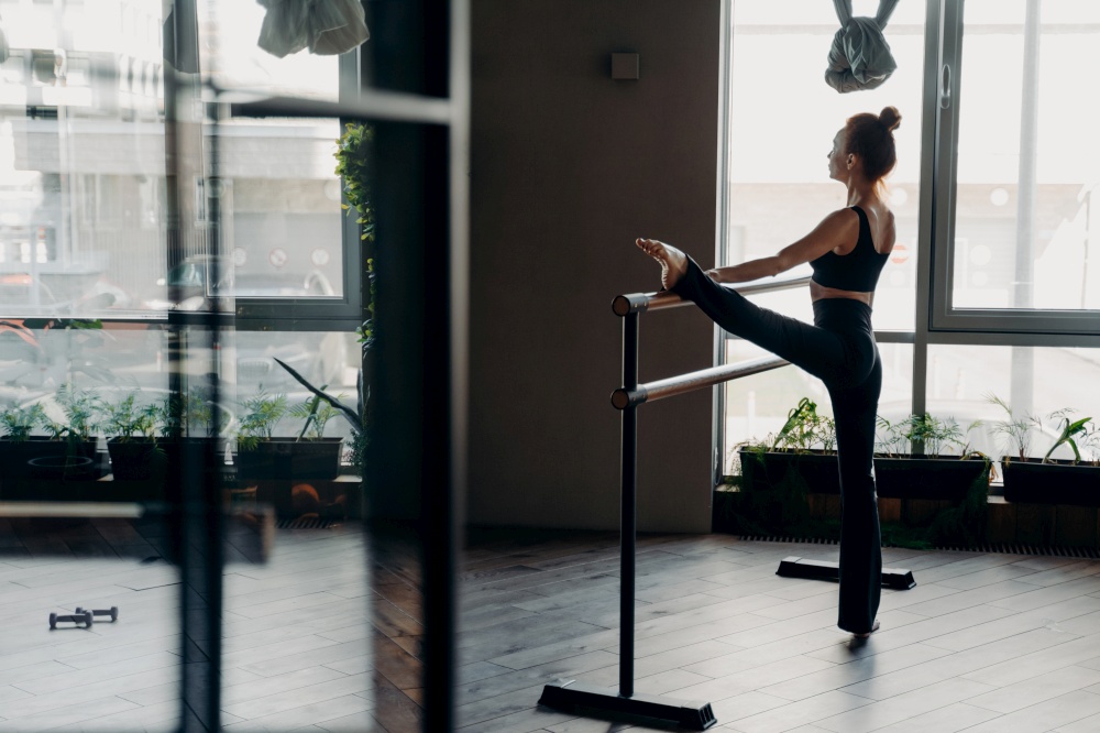 Slim red haired young ballerina in black top tank and leggings standing in split position next to ballet barre in gym against light large window background. Concept of stretching and sport indoor. Slim ballerina during her stretch routine next on ballet barre in studio