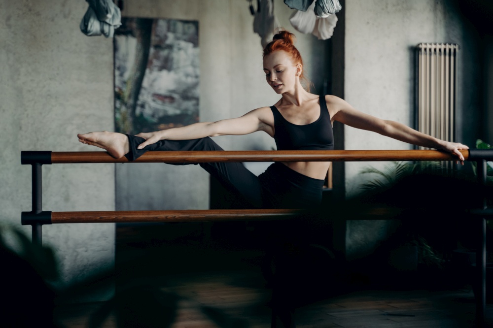 Working on flexibility. Young red haired woman ballerina lifted one leg on ballet barre and doing stretching exercises against darken background of studio or classroom. Barre workout concept. Young red haired woman in good body shape doing stretching exercises on ballet barre