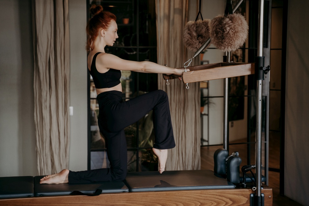 Side view of slender redhead female pilates trainer in black tight sportswear showing legs stretching exercises during training session in fitness studio, fit woman exercising in Cadillac reformer. Slender fit pilates trainer showing stretching exercises on cadillac reformer
