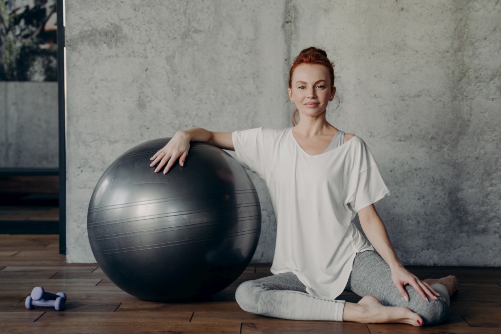 Portrait of beautiful sportive red haired lady gracefully sits on wooden floor in fitness or pilates studio and smiling happily at camera while leaning with one hand on fitball next to dumbbells. Beautiful sportive red haired lady gracefully sits on parquet floor with fitball