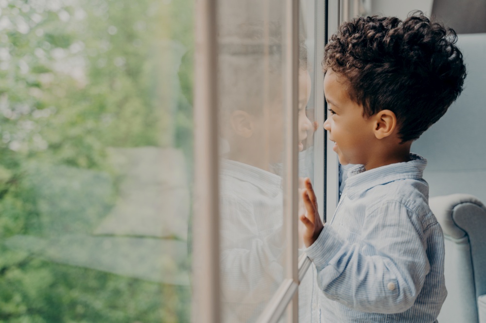 Side view of lovely afro american kid toddler waiting for mom, dressed in blue shirt, looking out of window through his reflection leaning on glass with hands and smiling. Children leisure time indoor. Lovely afro american kid in light blue shirt looking out of window