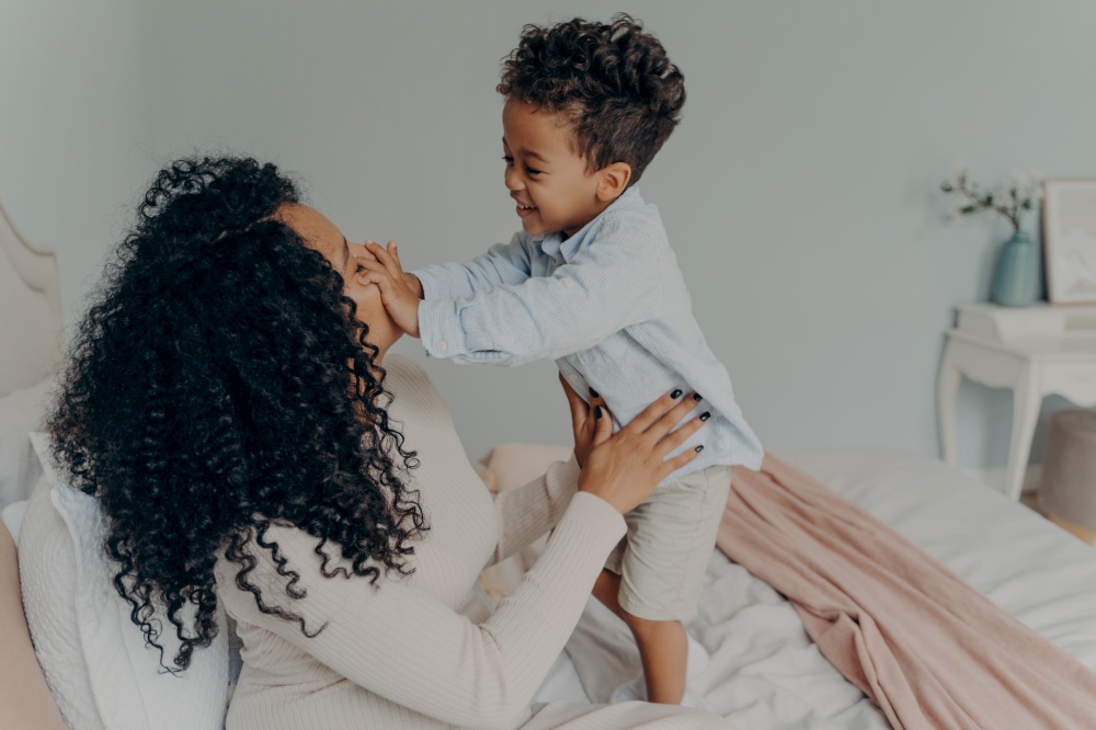 Happy afro american family, pregnant woman loving mom in white dress with curly hair sitting on white bed and joyfully spending time with child, giving attention to her son and playing with him. Pregnant afro american lady playing with her son at home