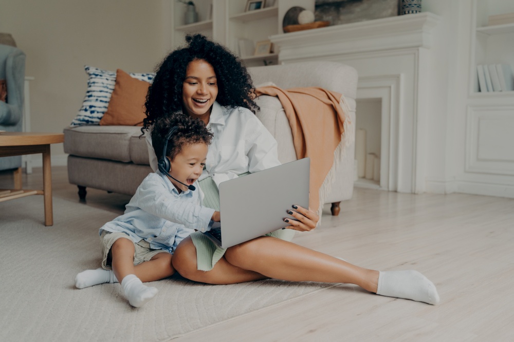 Young positive mixed race mother and cute boy son having video call with relatives, father or grandparents, sitting with laptop on floor and smiling, child wearing headset and pointing at screen. Young positive mixed race mother and cute boy son having video call on laptop