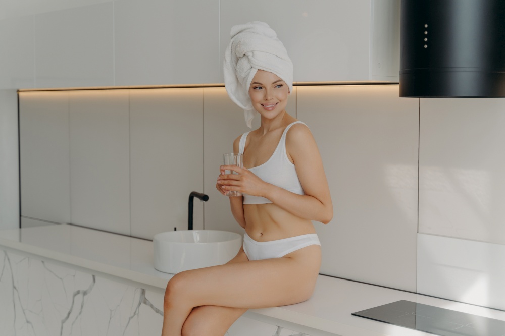 Healthcare concept. Fit slim female sitting on table top in modern stylish light colored kitchen and drinking mineral pure water from glass, looking fresh and clean after morning shower routine. Attractive fit young female sitting on marble table top with glass of water