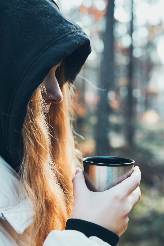 Woman in a hood having break during autumn trip holding cup with hot drink from thermos flask on autumn cold day. Active girl wandering in a forest actively spending time. Woman in a hood having break during autumn trip holding cup with hot drink from thermos flask on autumn cold day