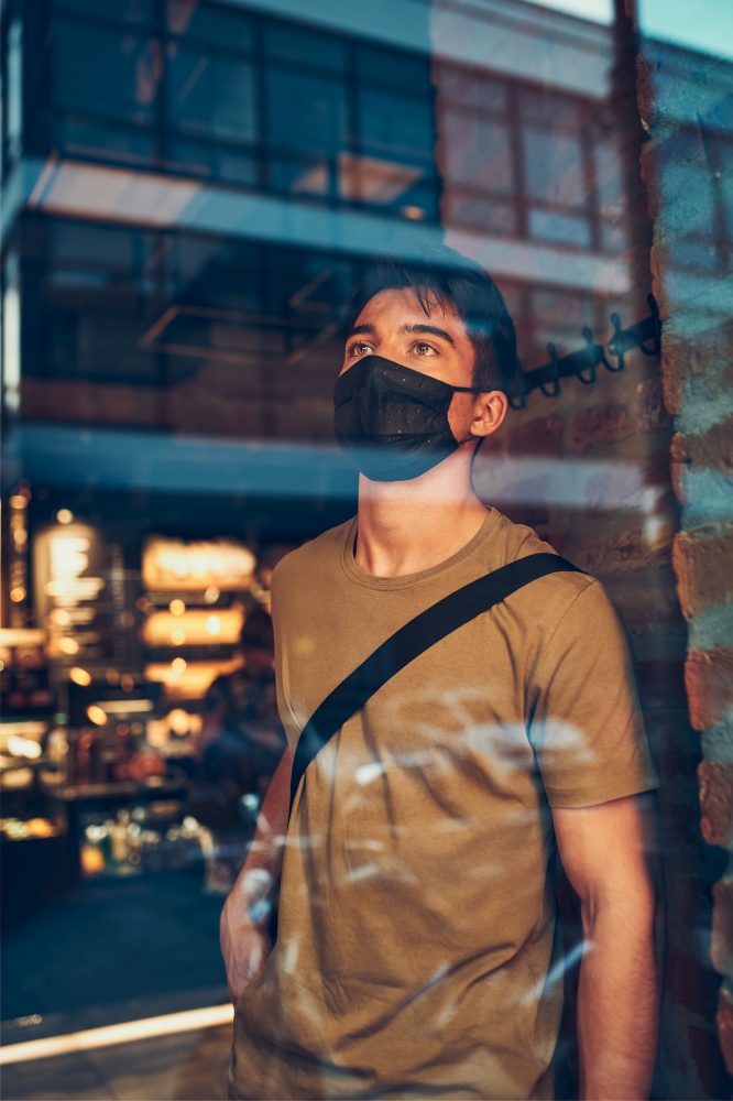 Young man standing in coffee shop at store front in the city center in the evening, wearing the face mask to avoid virus infection and to prevent the spread of disease in time of coronavirus